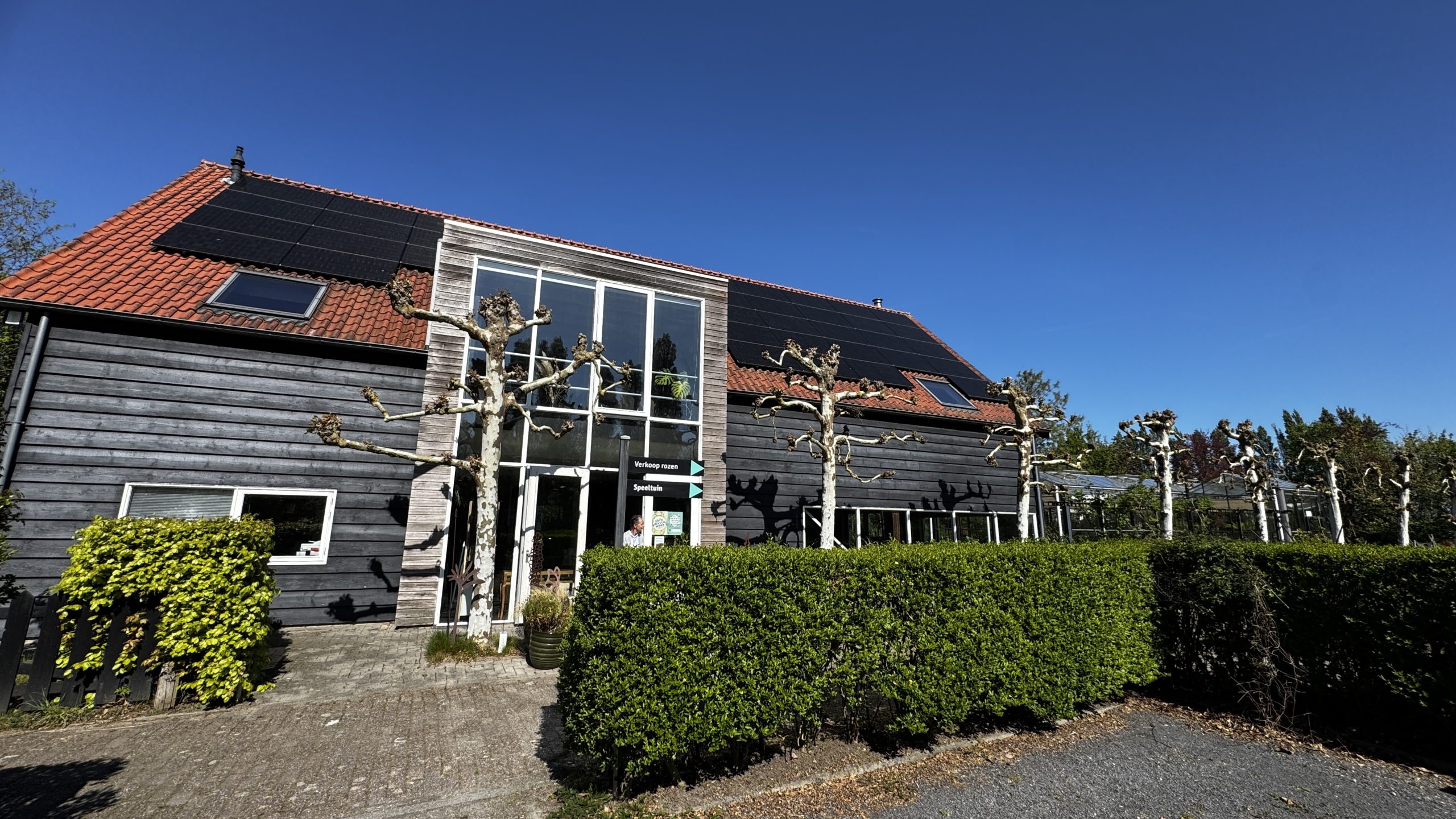 Two-story house with dark wooden siding and a red-tiled roof outfitted with solar panels, under a clear blue sky, with pruned trees and hedges in front.
