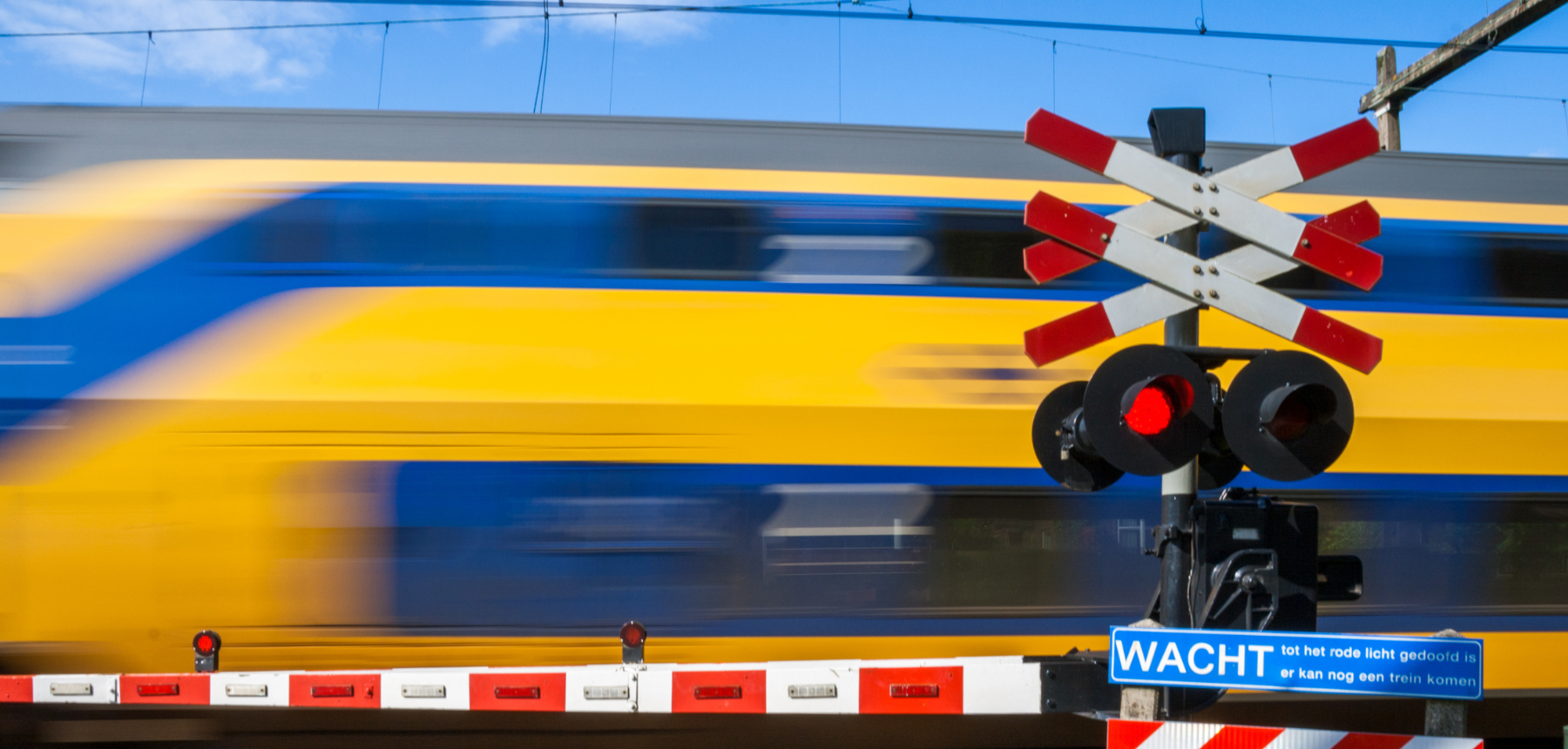 Blurred blue-and-yellow passenger train speeds by at a level crossing; red warning lights are illuminated and a blue 'WACHT' sign is visible.