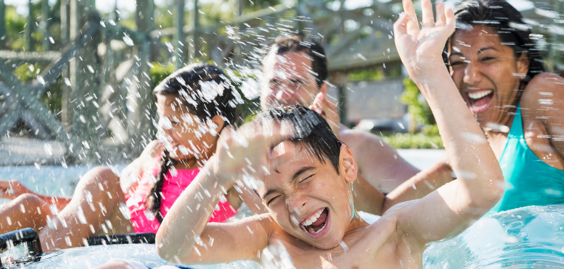 Group of kids and teens laughing and splashing water in a sunny outdoor pool.