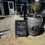 Outdoor café area with a chalkboard sign that says 'Welkom' and welcomes visitors; metal-and-wood barrel planter with a small palm tree, black patio furniture, and an open doorway in a dark wooden building.