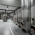 Interior of a beverage production facility with tall, polished stainless steel fermentation tanks and piping.