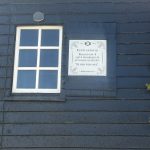 Blue wooden house exterior with a white-framed window and a decorative plaque mounted beside it on the wall.