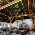 Rustic restaurant interior with exposed wooden beams, vaulted ceiling, and many tables set for dining under warm lighting.