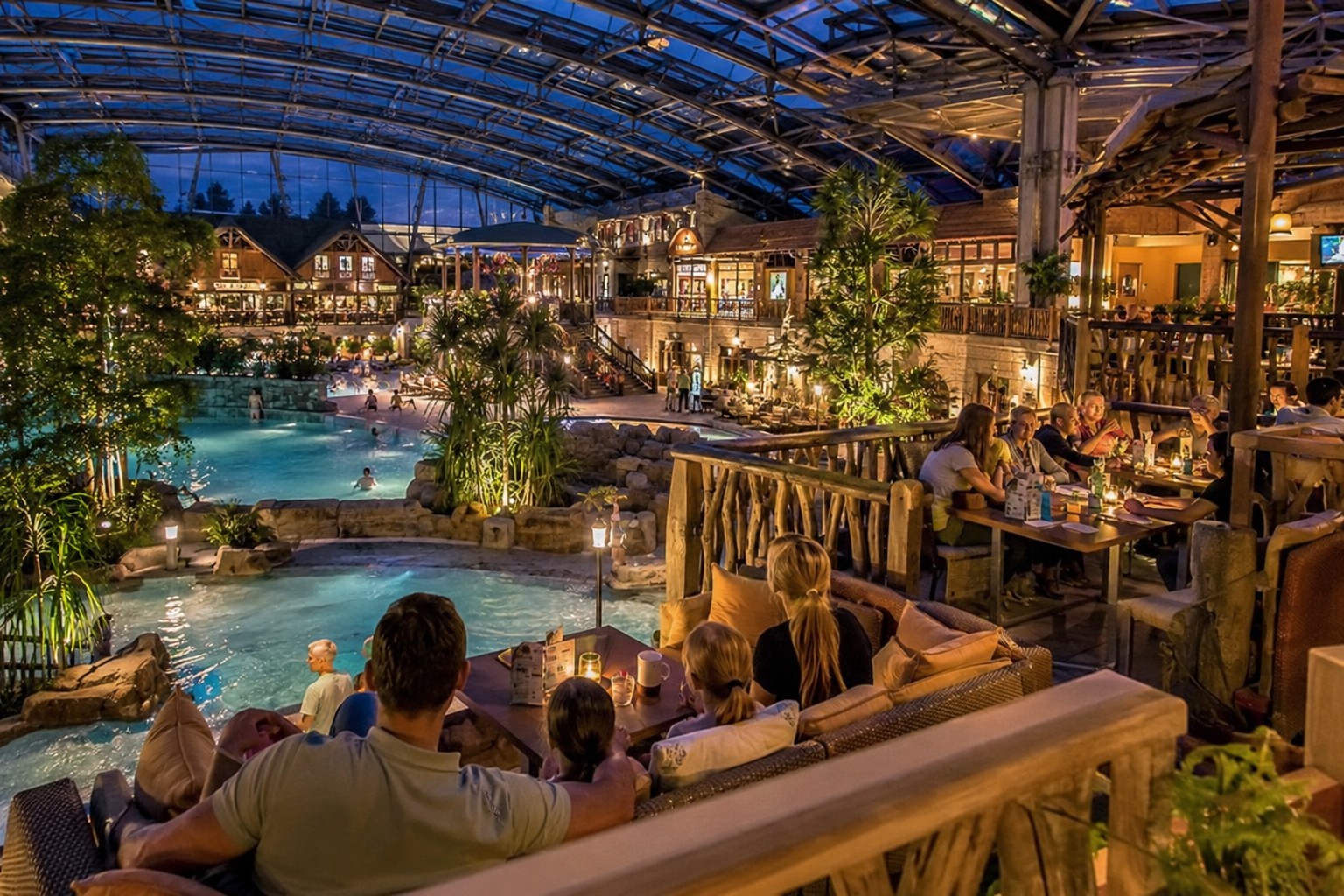 Indoor resort atrium with a central pool, tropical plants, and diners at wooden tables along a balcony grocery.