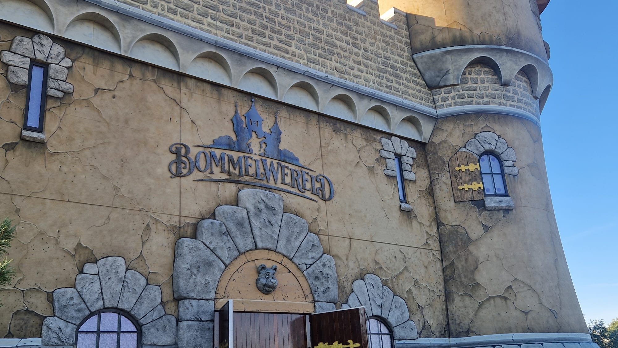 Fantasy castle-style exterior with arched stonework, small blue-framed windows, and a carved bear head above an arched doorway; weathered, beige wall.