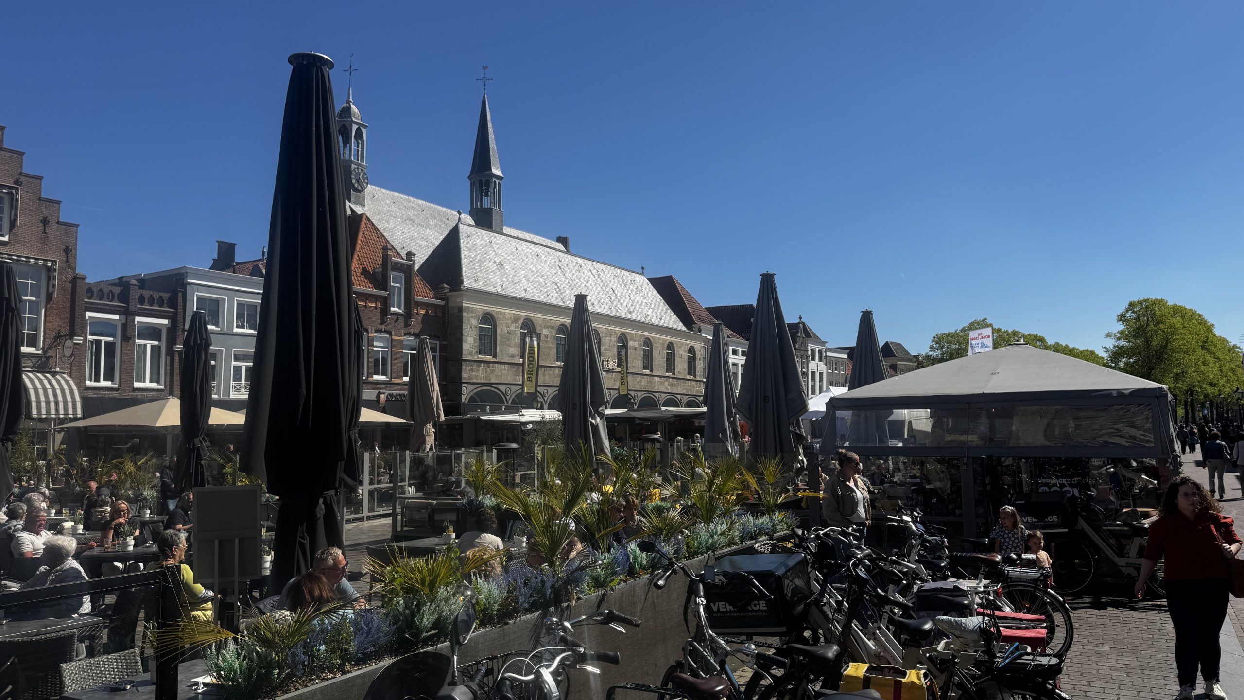 Outdoor cafe scene in a historic town square with dark umbrellas, diners at tables, and bicycles lined along a brick-paved street under a bright blue sky.