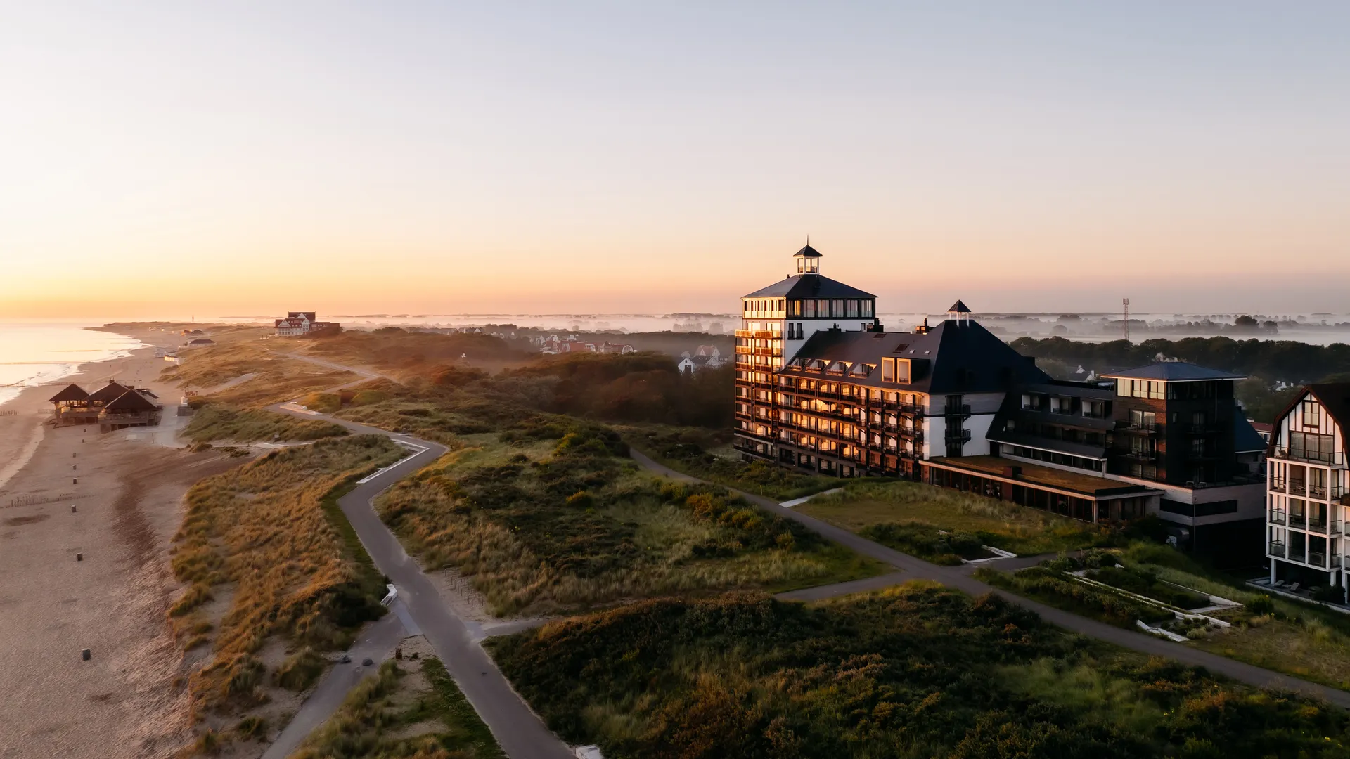 Sunset over a coastal resort with a large multi-story hotel by the dunes and a winding path along the beach.