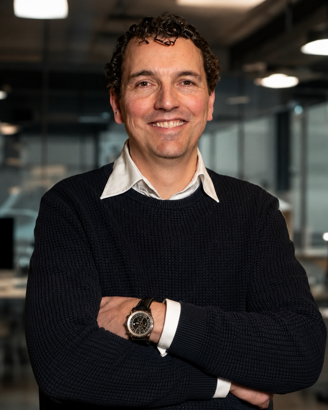 Smiling man with curly hair and a dark sweater, arms crossed in a modern office setting.
