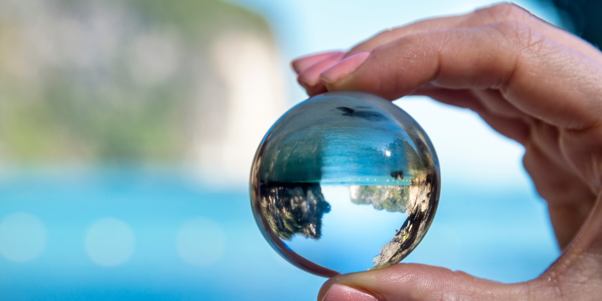 Close-up of a hand holding a glass marble; the sphere shows a reflected landscape and blue sky.